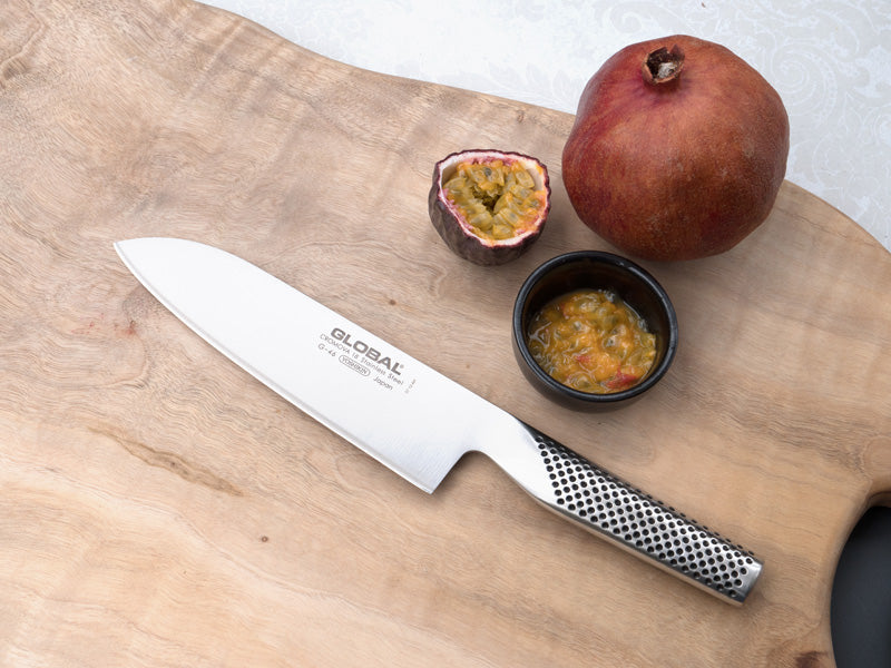 Ganzstahl Santokumesser von Global liegt auf Holzbrett neben einer Maracuja, einer Schüssel und einem Granatapfel.