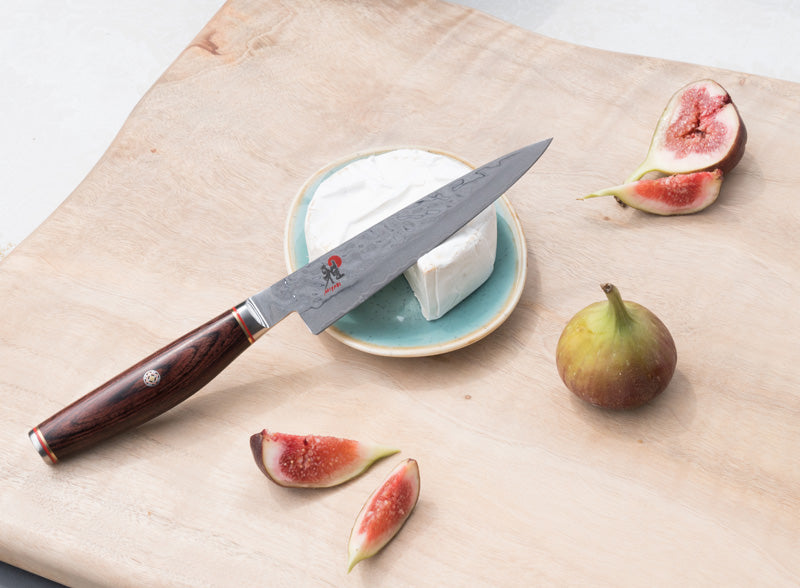 Ein Universalmesser von Miyabi. Die schmale kurze Klinge ist gehämmert und hochglanzpoliert. Der Griff ist leicht bauchig aus braunem Pakkaholz.
